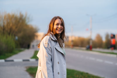 Beautiful smiling girl with long hair in a grey trench coat outdoors on the street spring