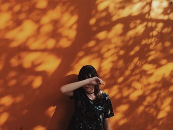 Woman hiding face with hand while standing against orange wall