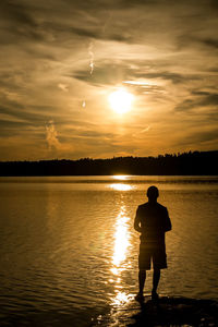 Rear view of silhouette man standing by lake against sky during sunset