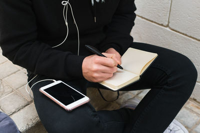 From above anonymous guy in casual clothes listening to music on smartphone and making notes in notepad while sitting on step on city street