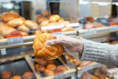 Gloved hand of mature female buyer holding fresh bun while standing by display in supermarket