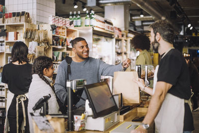 Male retail clerk giving shopping bag to customers at checkout in grocery store