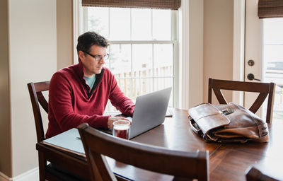 Man in glasses working from home using a computer at a dining table.