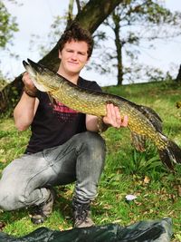 Young man holding fish while sitting at riverbank