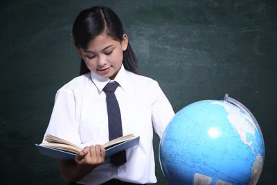 Schoolgirl reading book by globe against blackboard