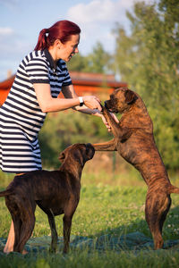 Mid adult woman playing with dogs while standing on grassy field against sky at park