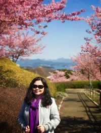 Portrait of smiling young woman standing by cherry tree against sky