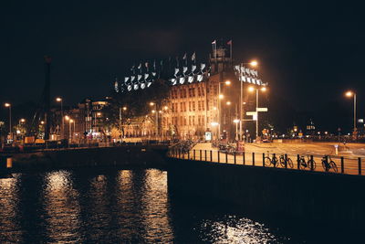 Illuminated buildings by river against sky at night