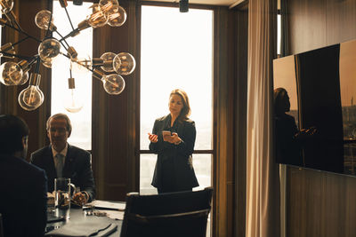 Confident female advisor talking on smart phone while standing by colleagues in board room at law office