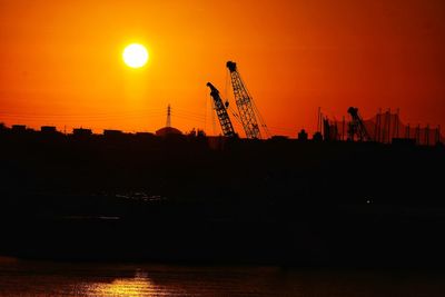 Silhouette cranes at construction site against orange sky