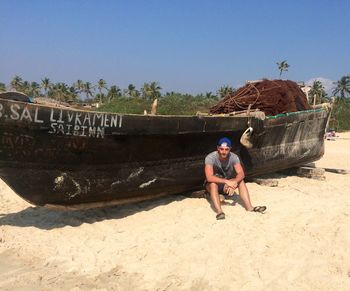 Young man sitting by old boat at beach against clear sky