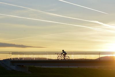 Man riding bicycle on bridge against sky during sunset