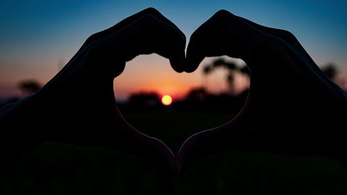Close-up of hand holding heart shape against sky during sunset
