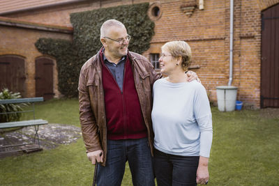 Smiling senior man with arm around woman standing against house at back yard