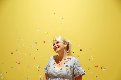 Confetti falling on cheerful young woman standing against yellow background