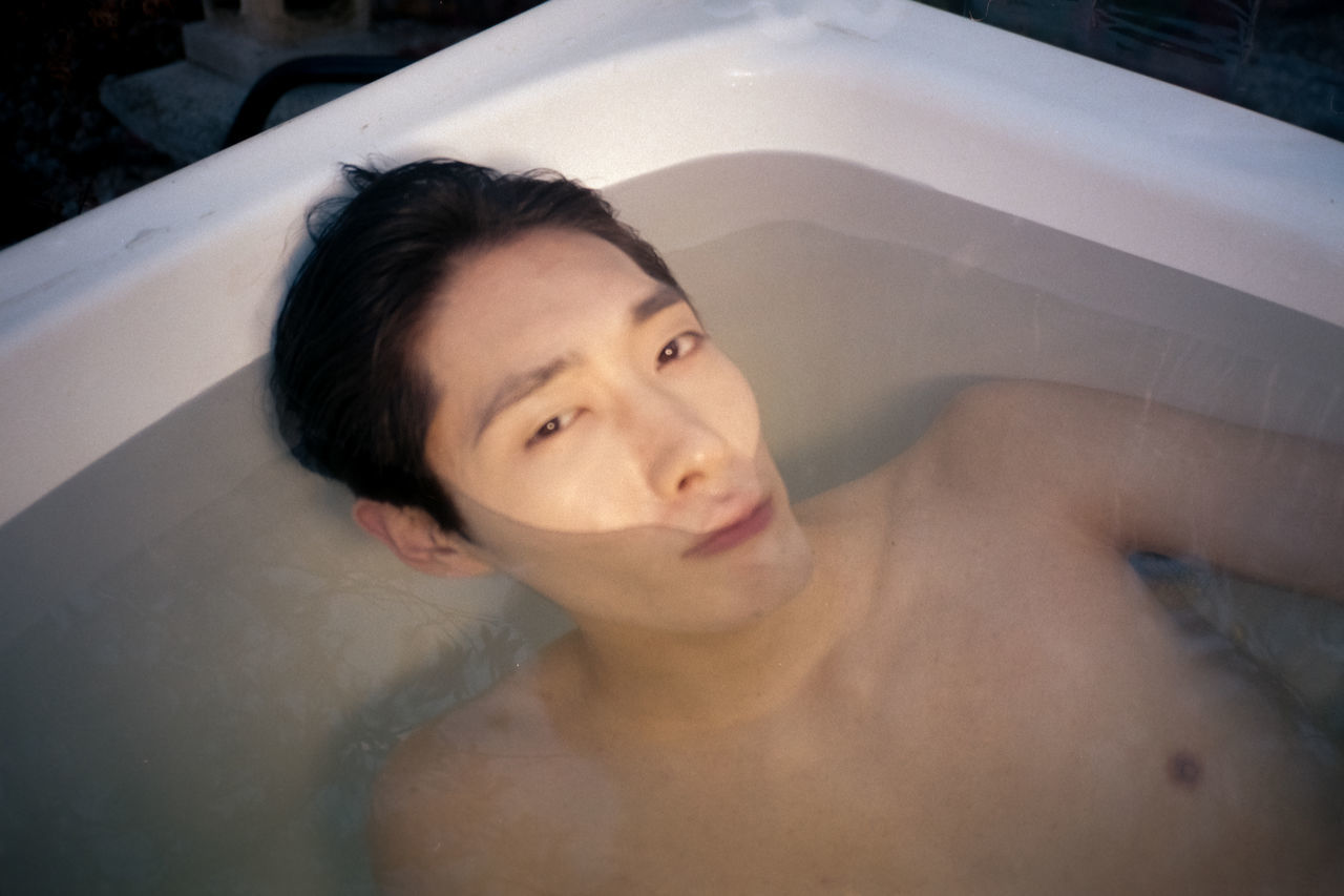 one person, young adult, headshot, portrait, front view, real people, leisure activity, lifestyles, shirtless, bathtub, looking at camera, adult, black hair, high angle view, young men, indoors, relaxation, looking