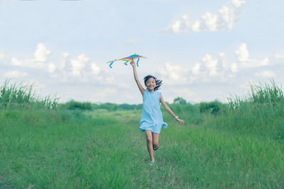 Full length of happy girl holding kite while running on grassy field against sky