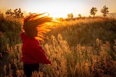 Side view of woman tossing hair while standing by plants against sky during sunset