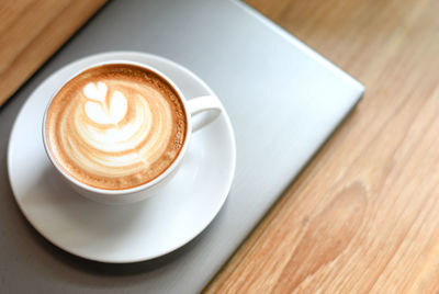 White latte coffee cup, decorated with leaves on the laptop placed on a wooden floor.