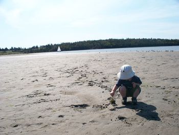 Boy with hat crouching at beach against sky on sunny day