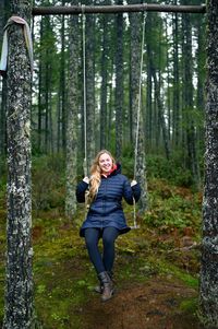 Portrait of smiling young woman swinging against trees in forest