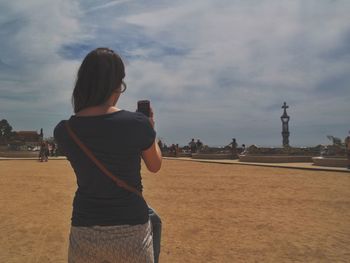 Rear view of woman photographing on mobile phone while standing on sand against cloudy sky