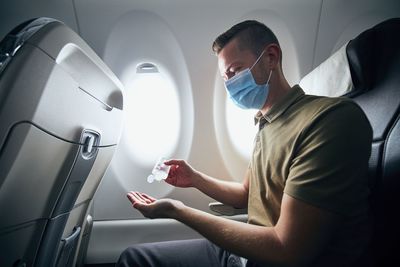 Man wearing face mask and using hand sanitizer inside airplane during flight.