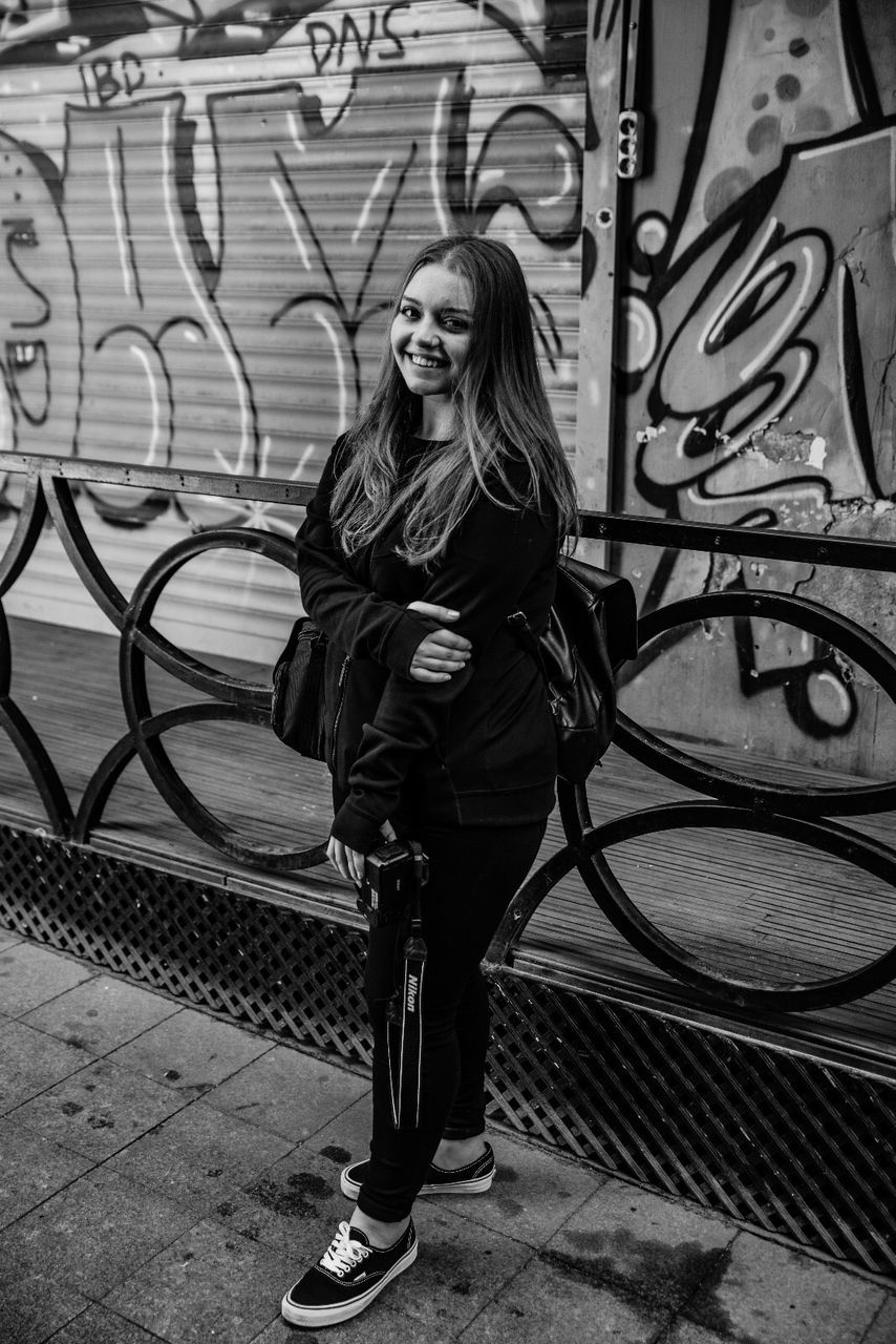 one person, black, full length, black and white, graffiti, standing, lifestyles, monochrome, young adult, women, monochrome photography, casual clothing, leisure activity, front view, street, architecture, adult, creativity, portrait, day, wall - building feature, white, art, city, built structure, fashion, clothing, road, footwear, hairstyle, long hair, looking at camera, looking, person, female, jacket, outdoors, street art, glasses, footpath, smiling, building exterior, drawing