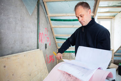 Serious manual worker reading blue prints while standing at construction site