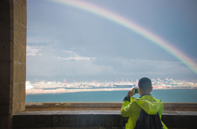 Rear view of man photographing standing by sea against rainbow in sky