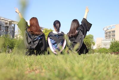 Rear view of friends wearing graduation gown while sitting on grass