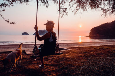 Side view of senior woman sitting on swing at beach against sky during sunset