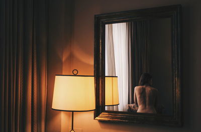 Rear view of shirtless woman reflecting on mirror by illuminated lamp at home