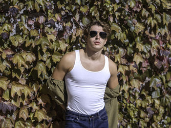 Stylish guy in sunglasses and casual clothes standing against ivy with autumn leaves