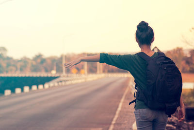 Rear view of female hitchhiker standing on roadside against sky