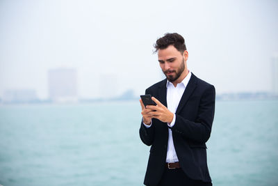 Businessman using phone while standing at beach against clear sky