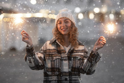 Portrait of young woman holding sparkler outdoors during winter