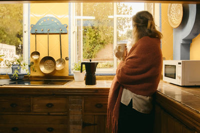 Senior woman in covered blanket looking through window while drinking coffee at home