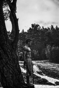 Side view of shirtless man standing by tree against sky