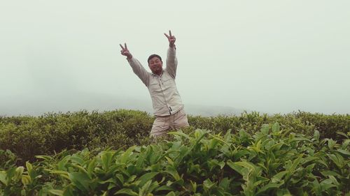 Portrait of man with arms raised showing peace sign while standing by plants on land against sky