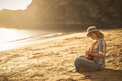 Woman in sunglasses playing guitar while sitting at beach