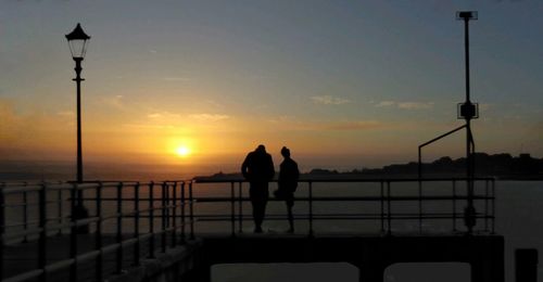 Silhouette people on street by sea against sky during sunset