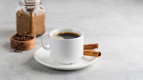 Black coffee in a white mug with cinnamon sticks on a saucer on a grey table. copy space.