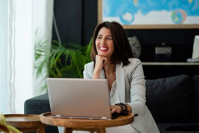 Thoughtful smiling woman using laptop while sitting on sofa at home