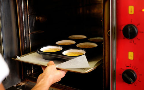 Crop anonymous pastry chef putting muffin tray with dough into industrial oven while preparing dessert in professional kitchen