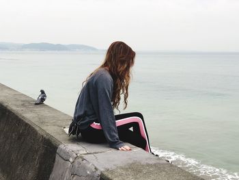 Woman sitting on retaining wall while looking at sea against sky