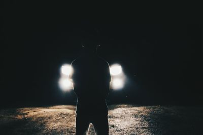 Rear view of silhouette man standing by illuminated car headlights at night