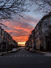 Cars on road by buildings against sky during sunset