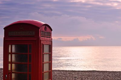 Red telephone booth by sea against sky during sunset