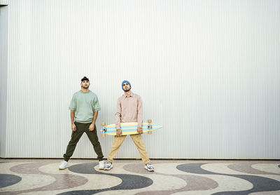 Two men with longboard standing in front of white wall